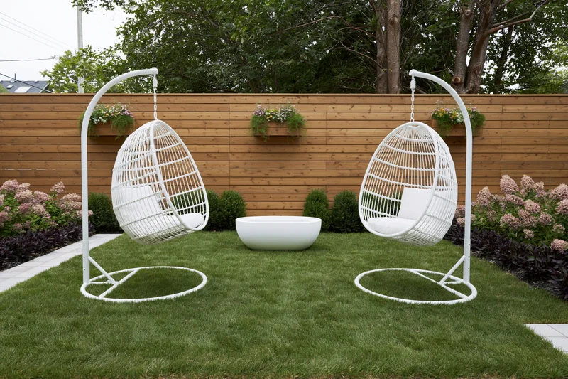 Two white rattan hanging egg chairs on freestanding frames in a grassy backyard garden with wooden fence and flowering shrubs