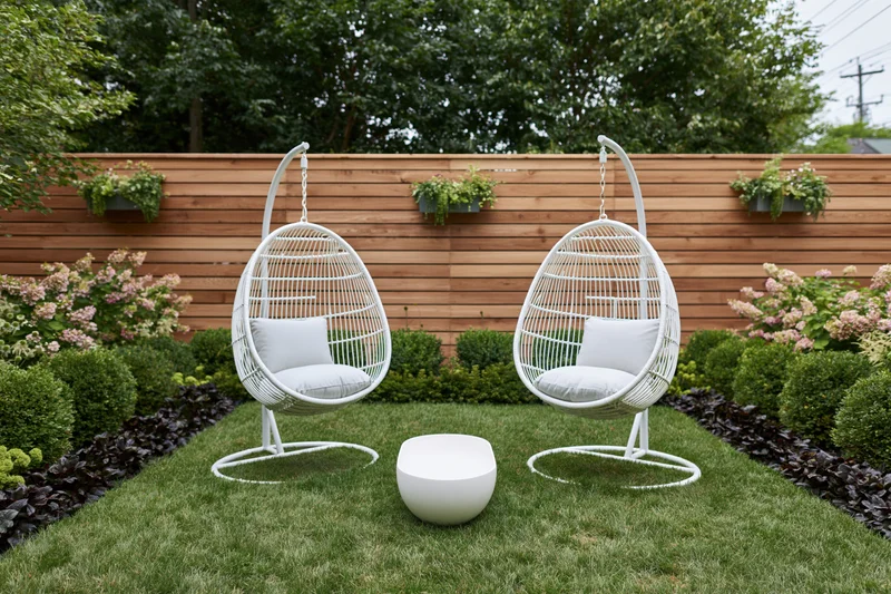 Two white rattan hanging egg chairs on freestanding frames in a landscaped backyard garden with green grass, shrubs, flowers, and a wooden fence