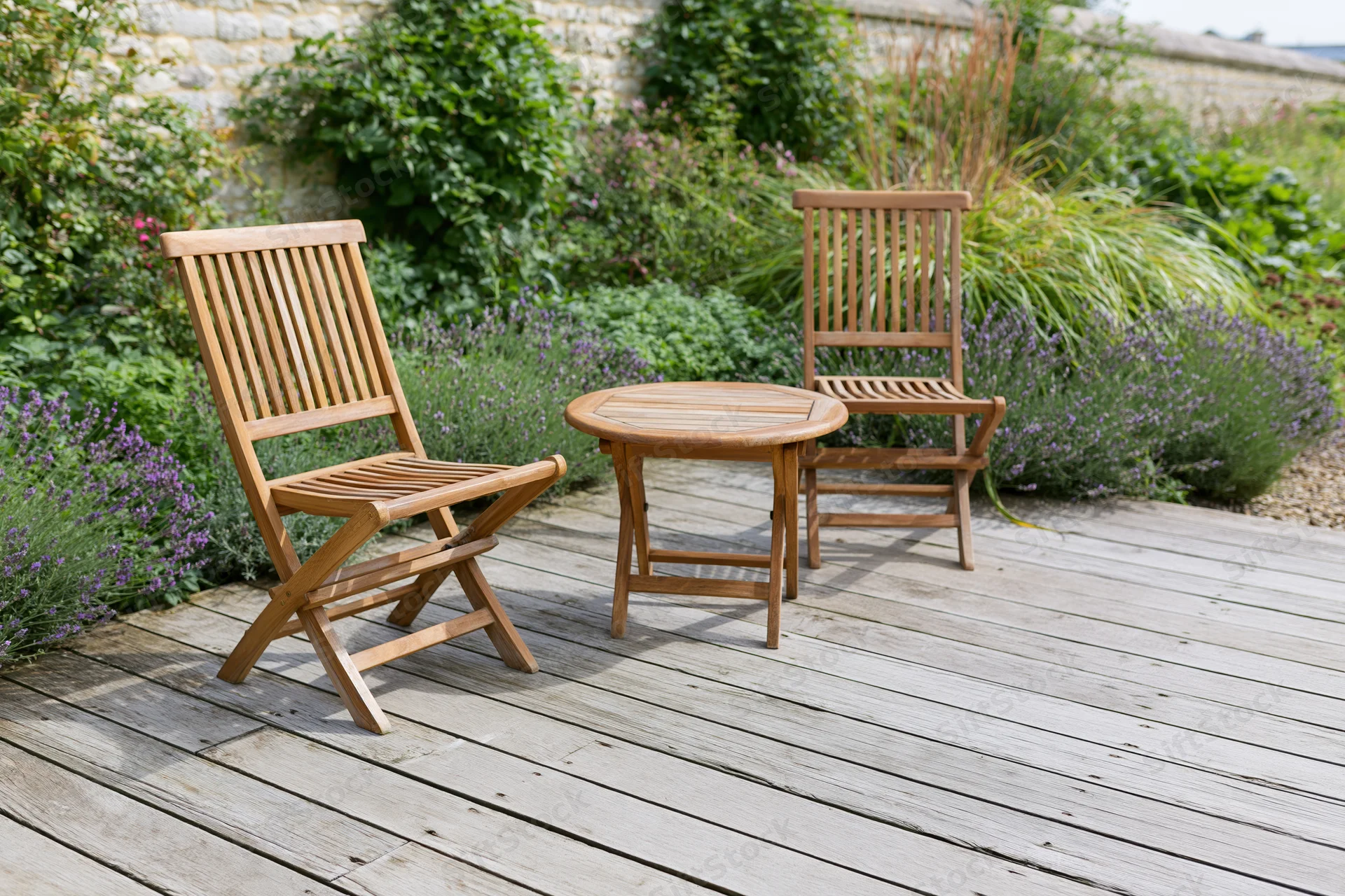 Two wooden folding chairs and a small round table on a weathered wooden deck surrounded by lavender and greenery