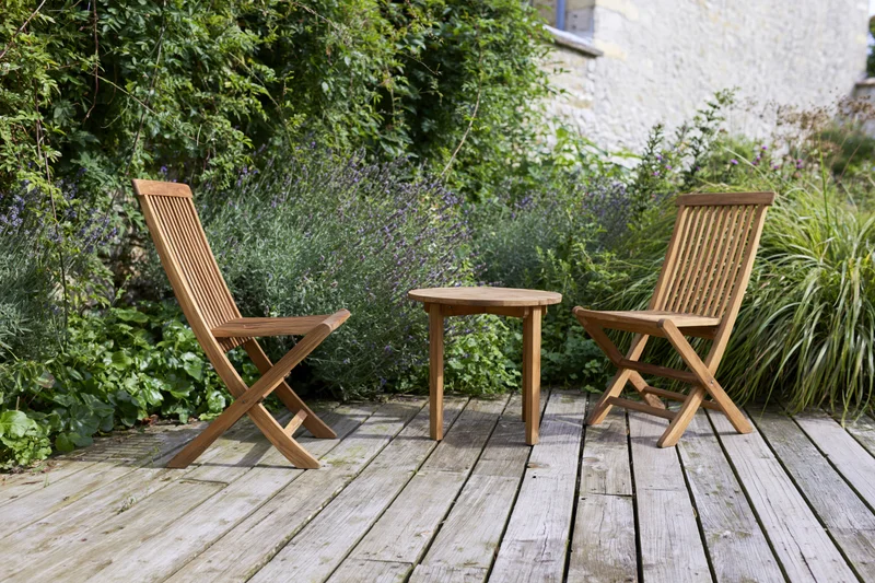 Two wooden folding chairs and a small round table on a weathered deck surrounded by greenery and lavender plants