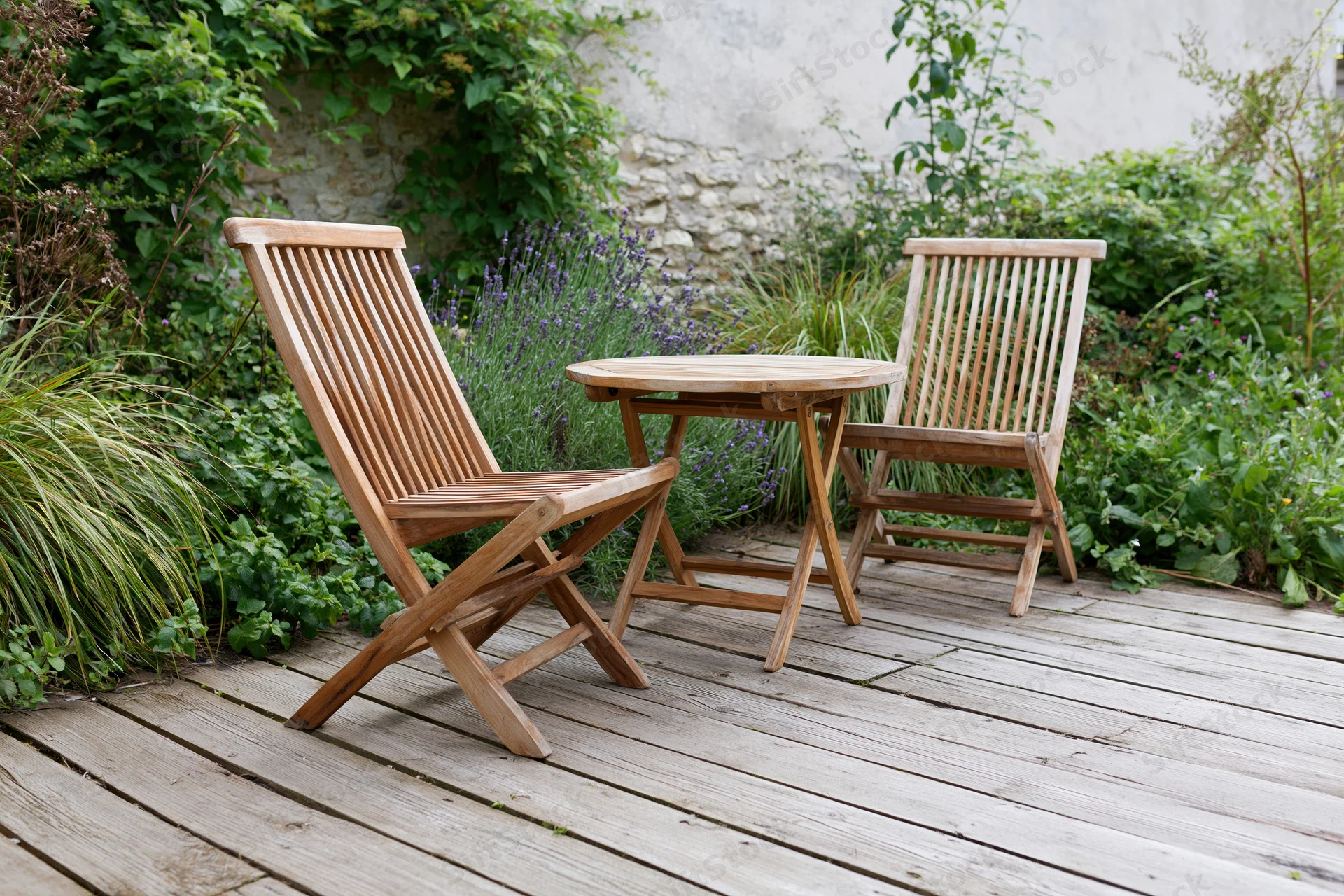 Two wooden folding chairs and a small round table on a weathered deck surrounded by greenery and lavender plants