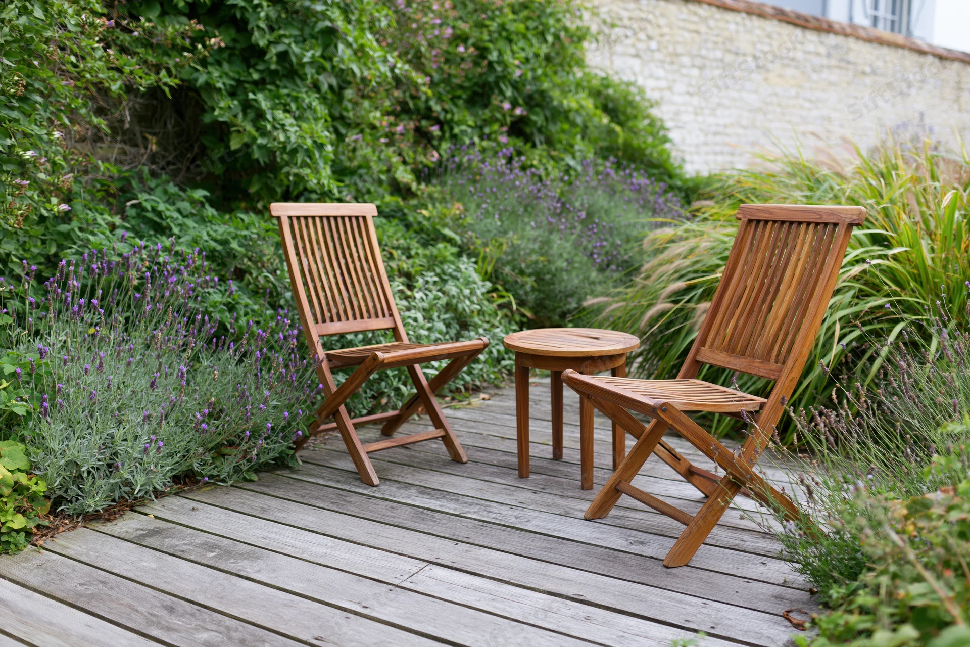 Two wooden folding chairs and a small round table on a deck surrounded by lavender and greenery