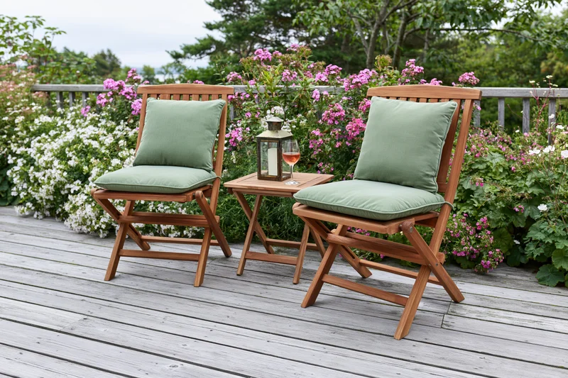 Two wooden folding chairs with green cushions on a deck surrounded by flowering shrubs and a small table with a lantern and wine glass