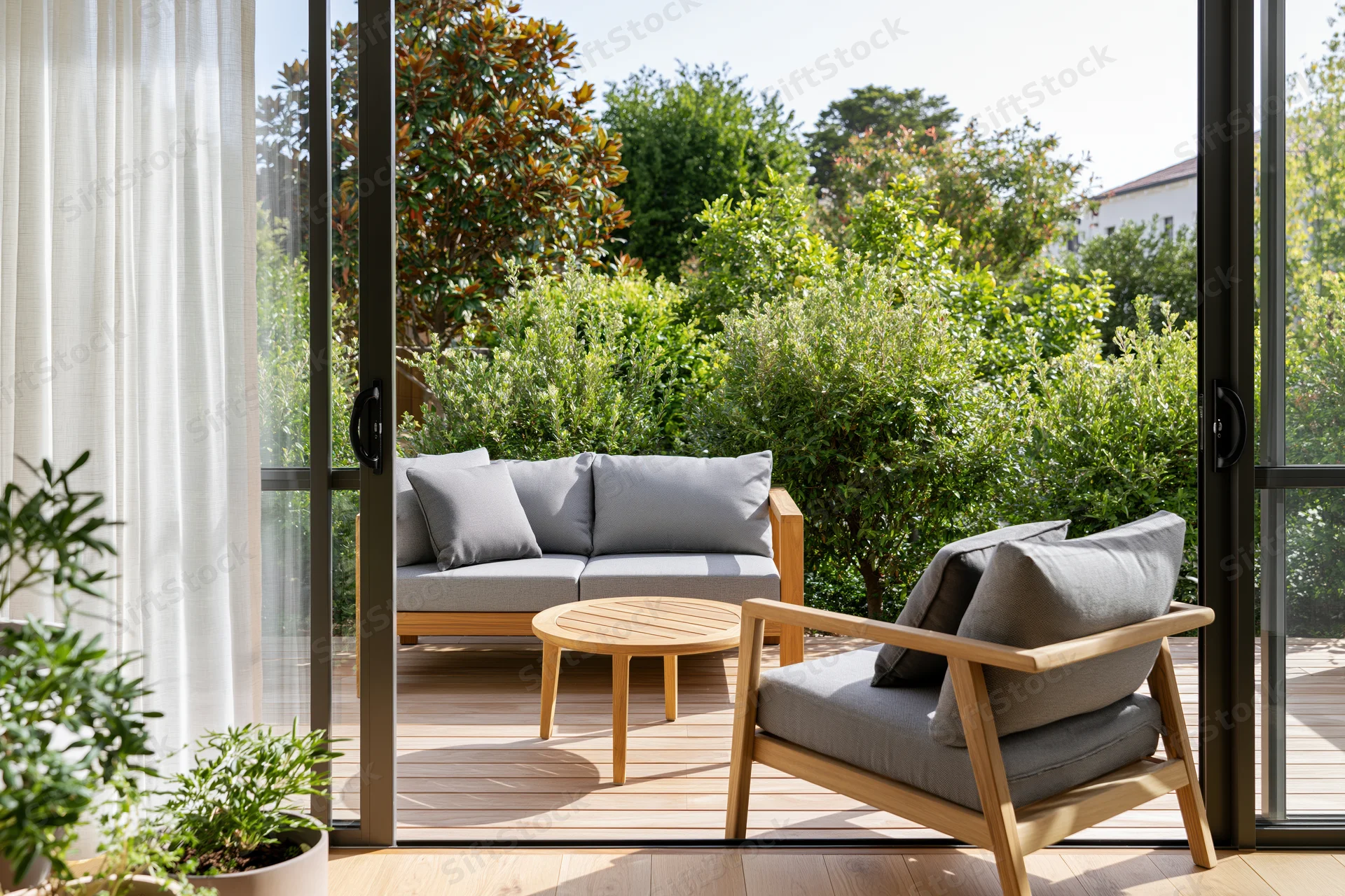 View through open glass sliding doors onto a sunny outdoor patio with wooden furniture and greenery