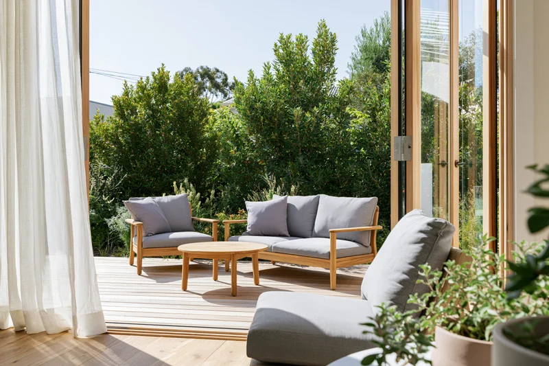 View through open glass sliding doors onto a sunny outdoor patio with wooden furniture and greenery
