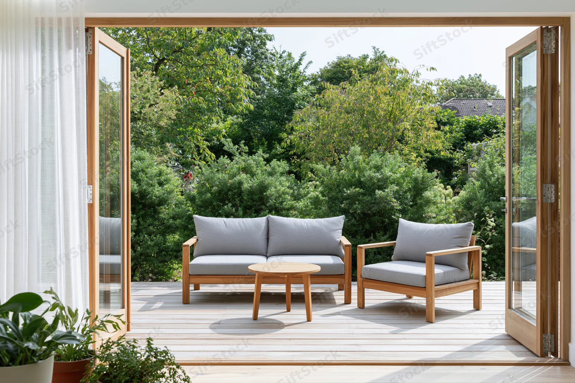 View through open glass doors onto a sunny patio with wooden furniture and greenery