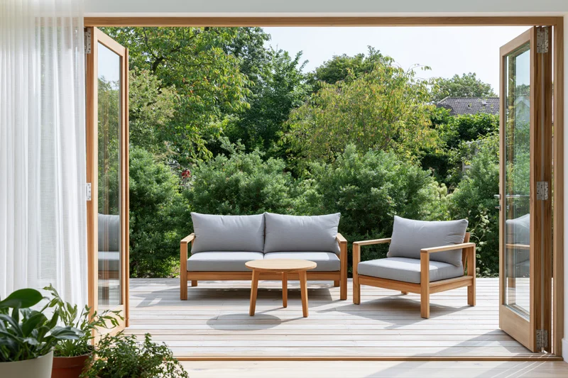 View through open glass doors onto a sunny patio with wooden furniture and greenery