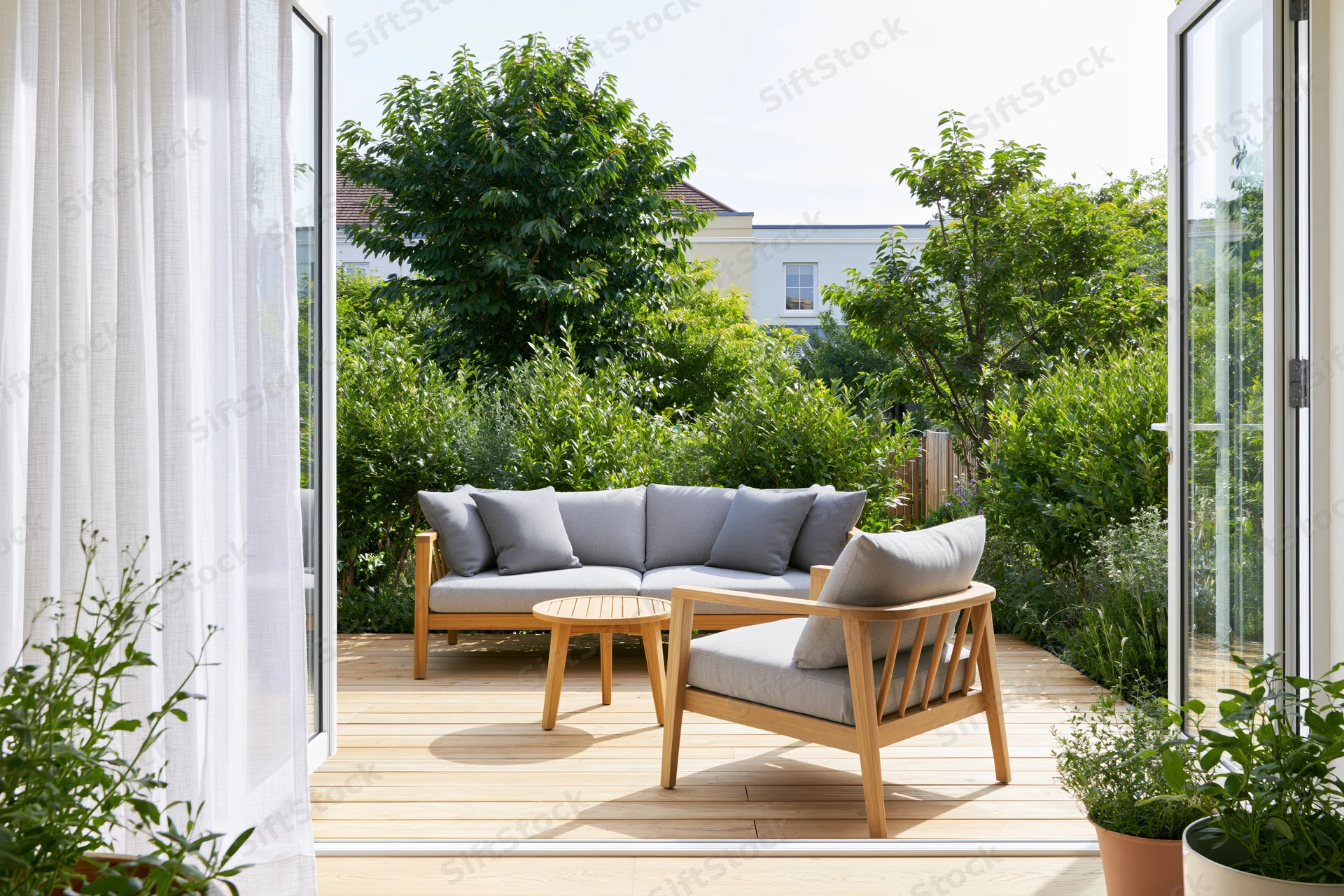 View through open glass sliding doors onto a sunny outdoor patio with wooden furniture and greenery
