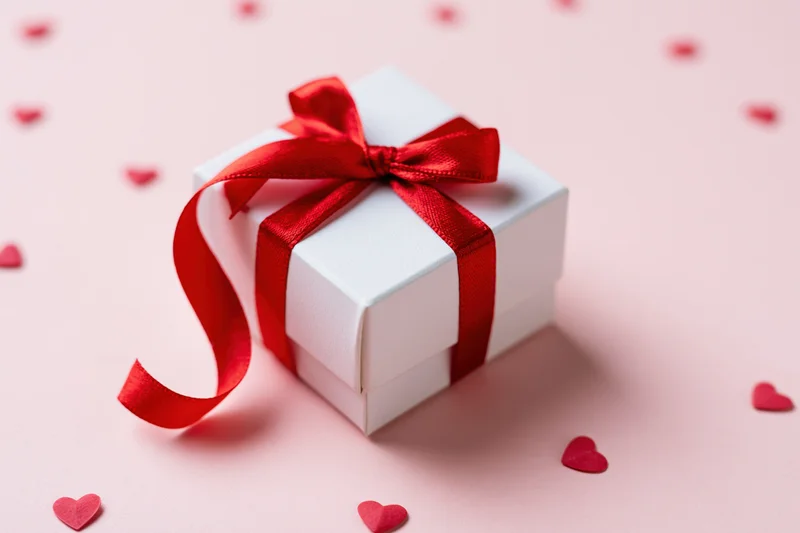 White gift box tied with red satin ribbon and bow on pink surface with scattered red hearts