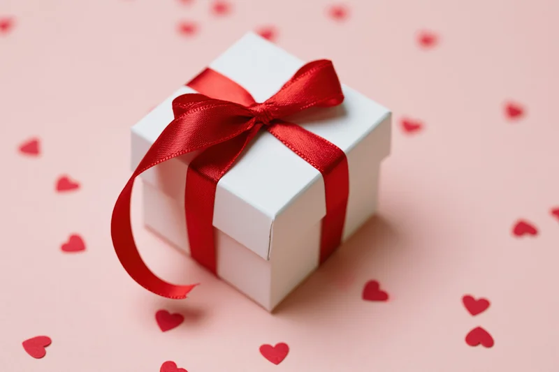 White gift box tied with red satin ribbon and bow on pink surface with scattered red hearts
