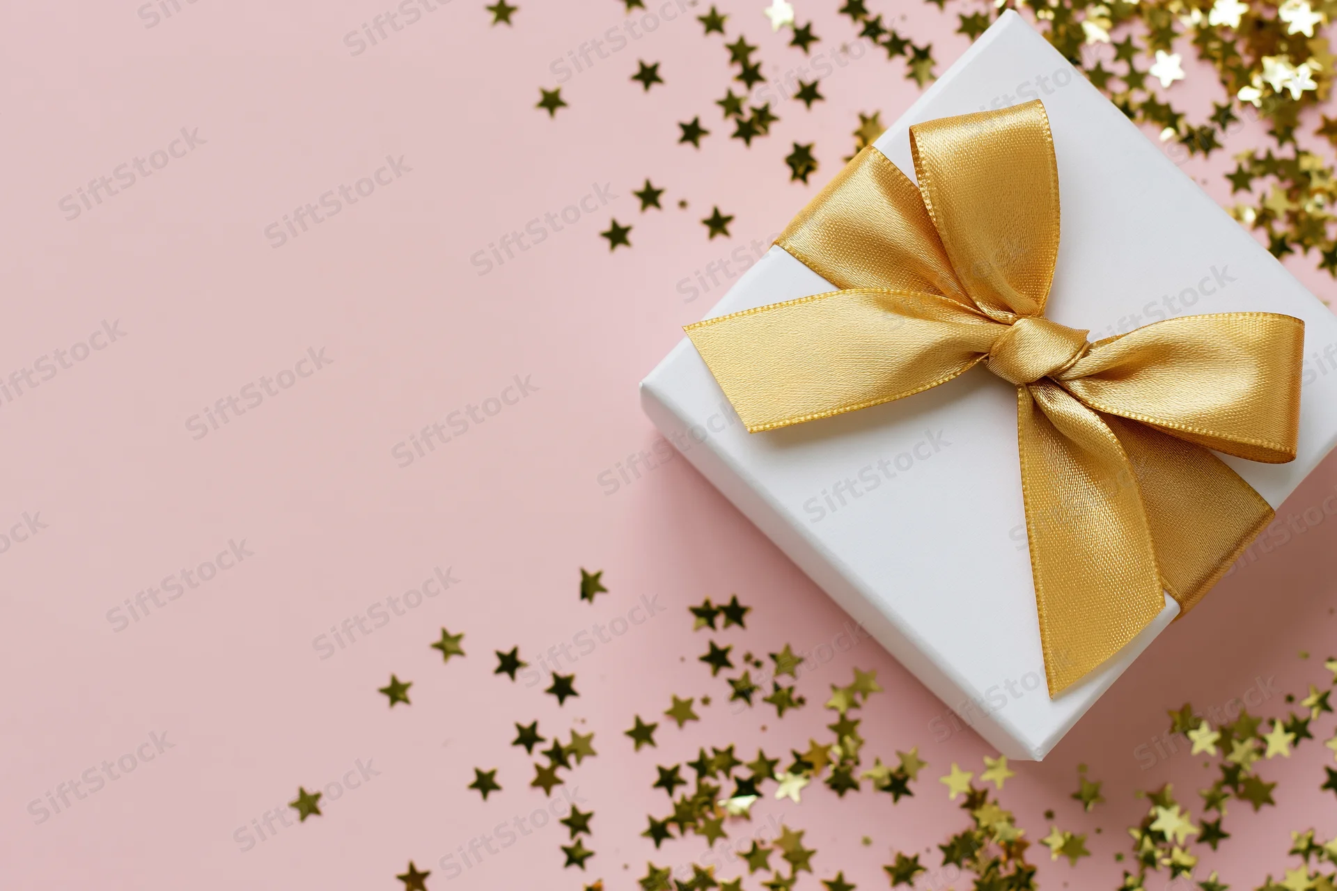 White gift box with gold satin ribbon bow on pink surface with star confetti