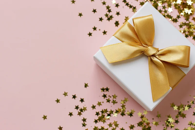White gift box with gold satin ribbon bow on pink surface with star confetti