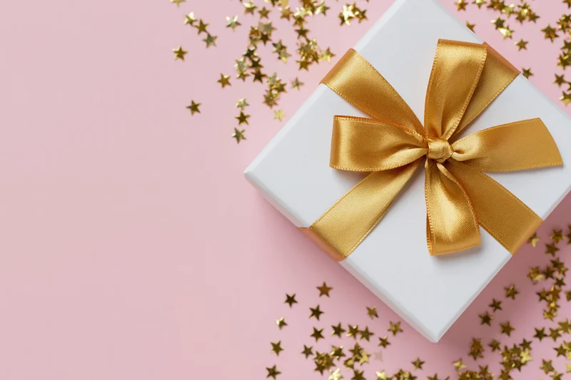 White gift box with gold satin ribbon bow on pink surface surrounded by star confetti