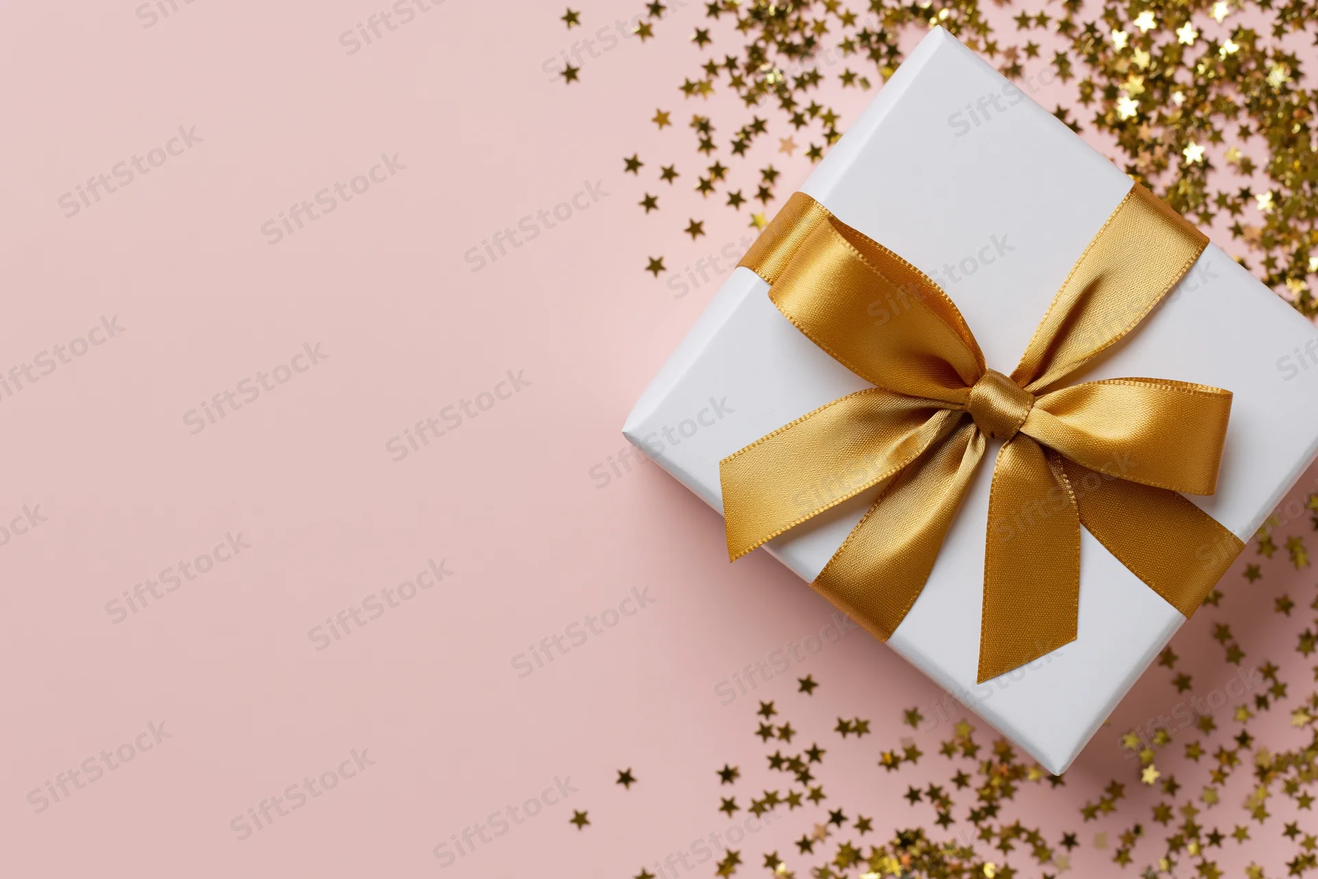 White gift box with gold satin ribbon bow on pink surface surrounded by star-shaped glitter confetti