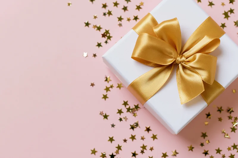 White gift box with gold satin ribbon bow on pink surface surrounded by star confetti