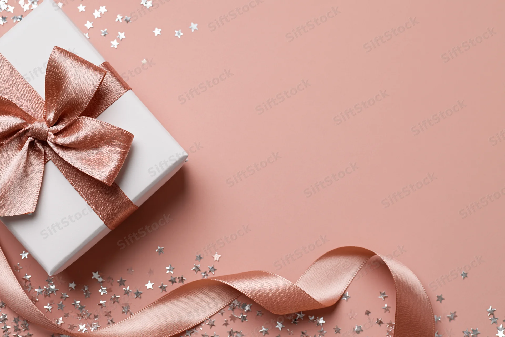 White gift box with rose-gold satin bow and silver star confetti on pink surface