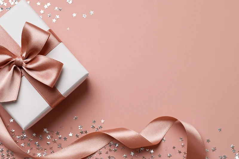 White gift box with rose-gold satin bow and silver star confetti on pink surface