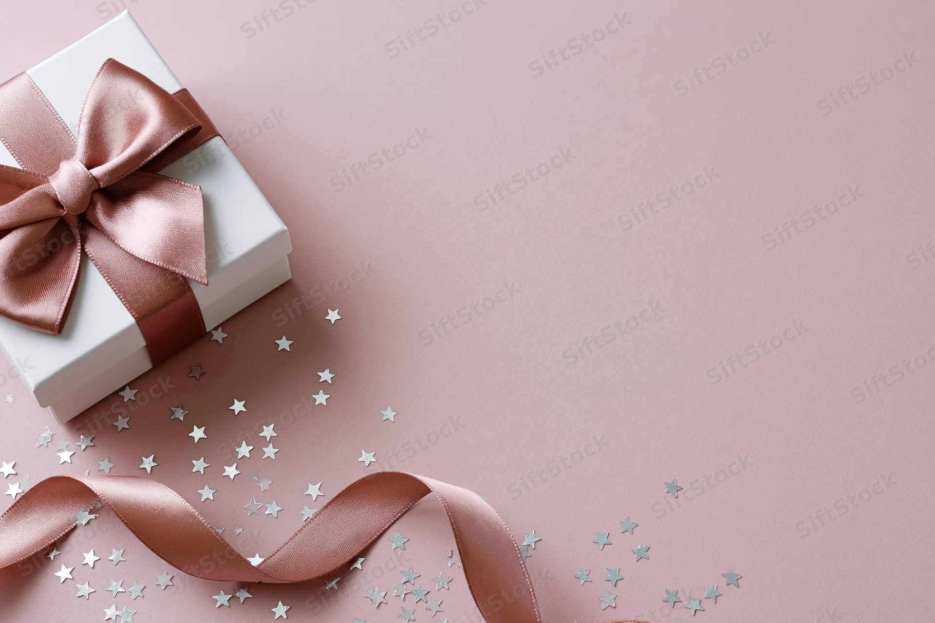 White gift box with rose gold satin bow and scattered star confetti on pink surface