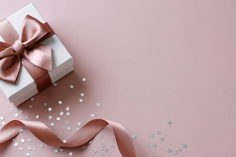 White gift box with rose gold satin bow and scattered star confetti on pink surface