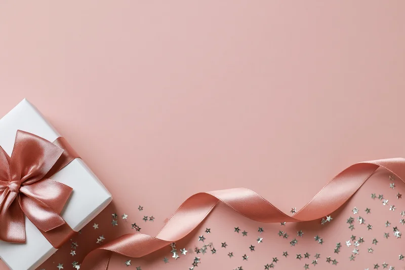 White gift box with rose-gold satin bow and scattered silver stars on pink background