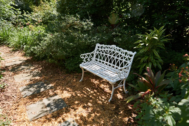 White ornate cast-iron bench on mulched garden path surrounded by greenery
