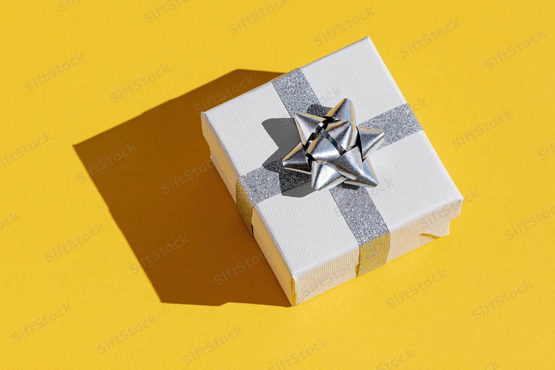White gift box with silver glitter ribbon and star bow on yellow background