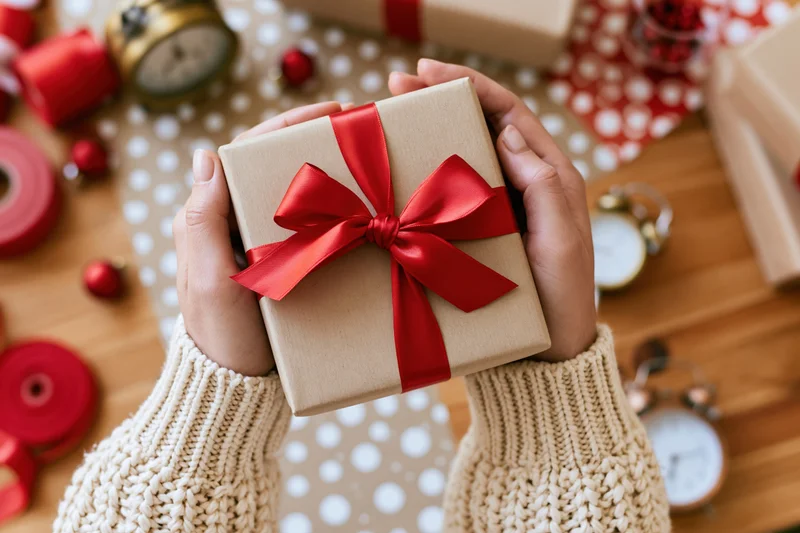 Woman's hands holding a kraft paper gift box with red ribbon bow over a decorated wooden table
