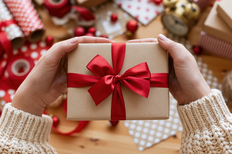 Woman's hands holding a kraft paper gift box tied with a red ribbon over a wooden table with wrapping supplies