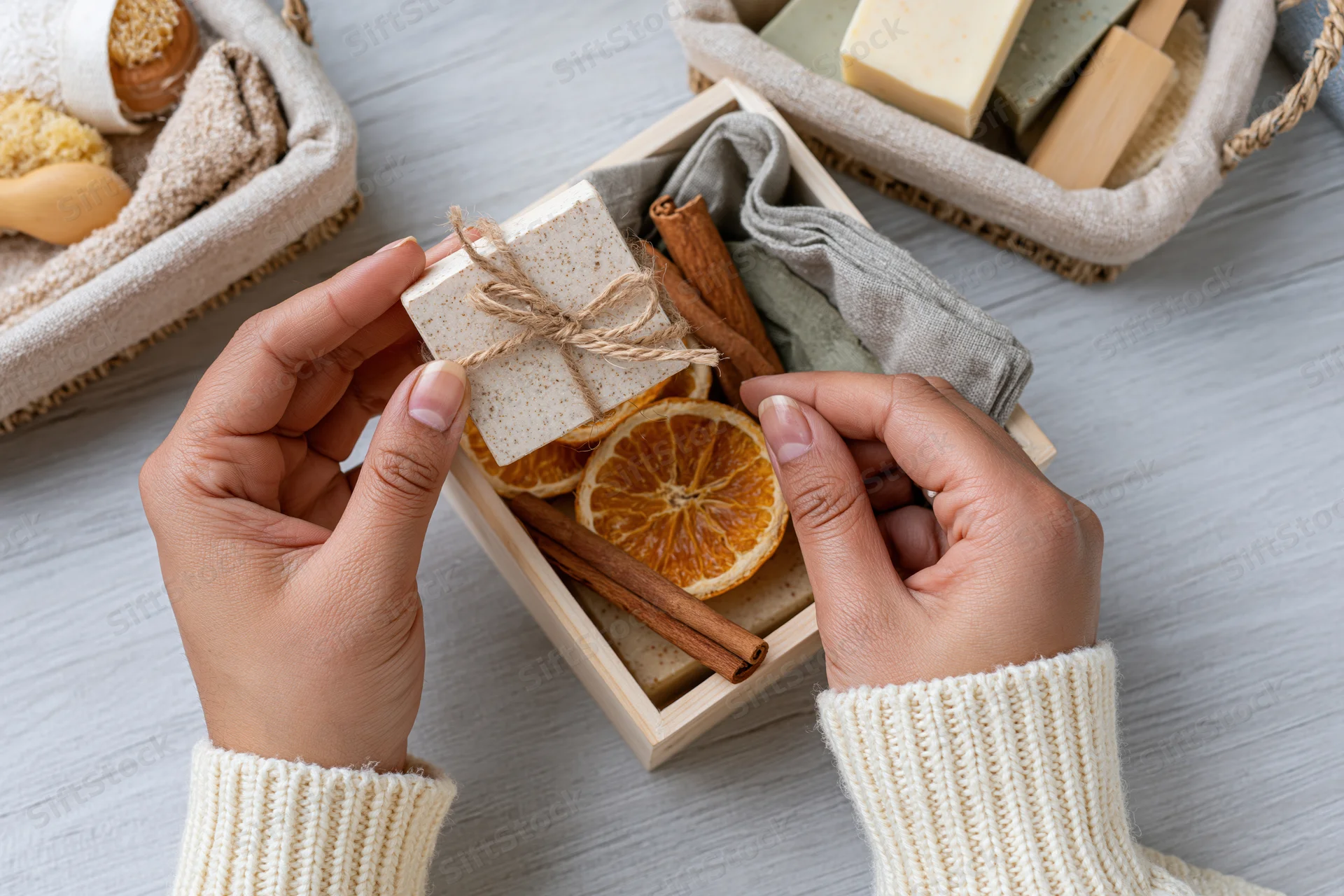 Woman's hands holding a handmade soap bar tied with twine over a wooden box filled with dried orange slices and cinnamon sticks on a light wood surface