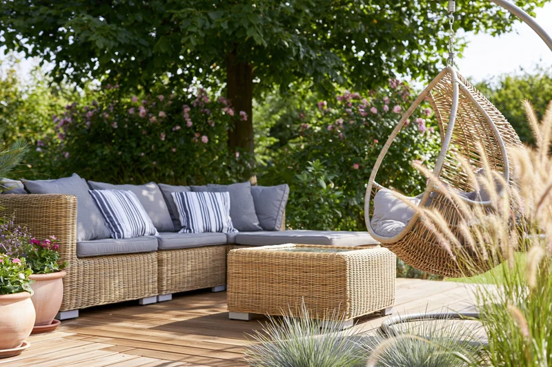 Rattan sectional sofa with hanging chair on wooden deck patio surrounded by greenery and flowers