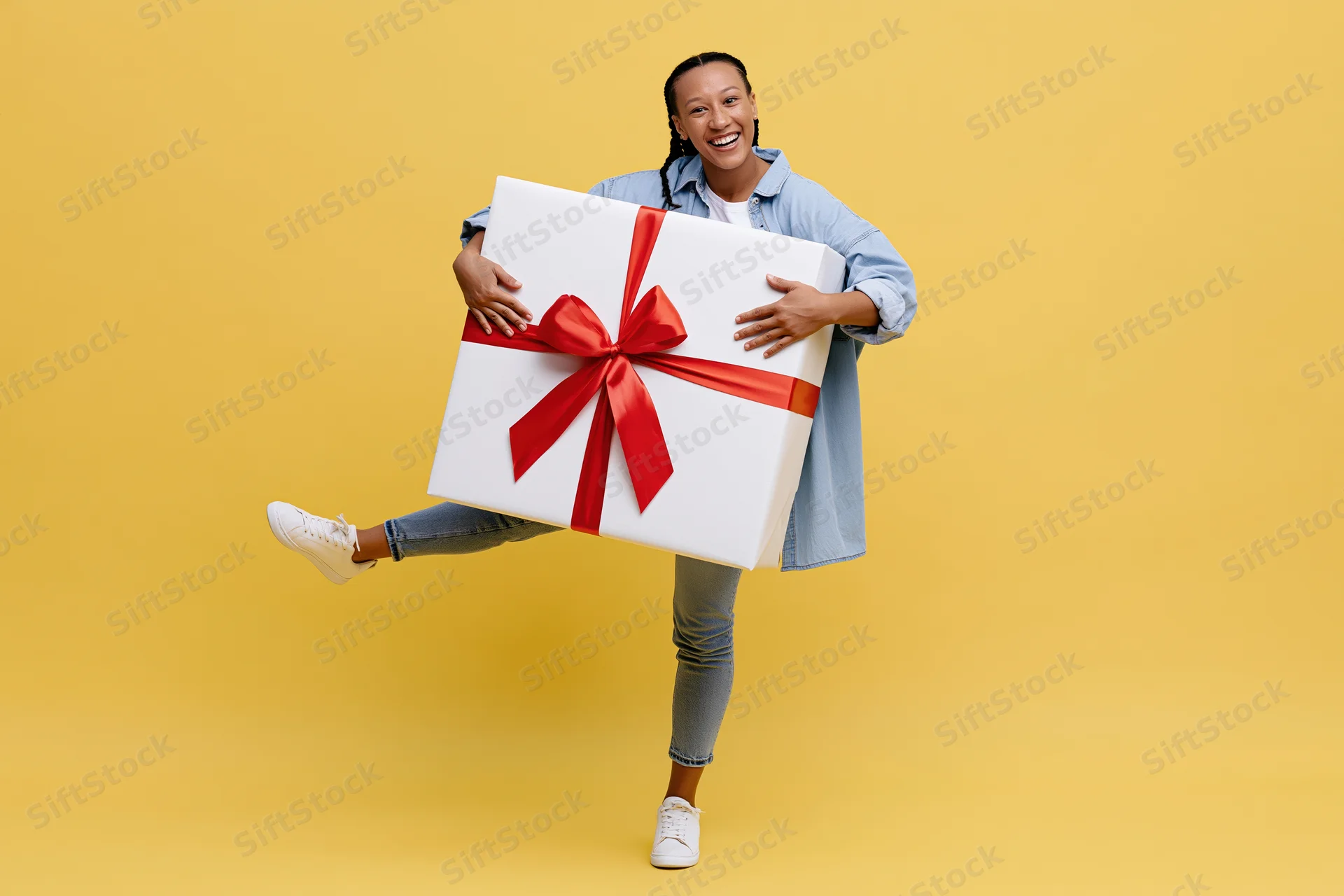 Woman smiling while holding oversized white gift box with red ribbon against yellow background