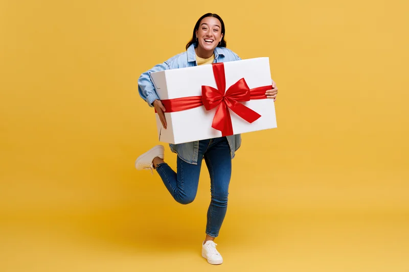 Woman smiling and jumping while holding a large white gift box with red ribbon against yellow background