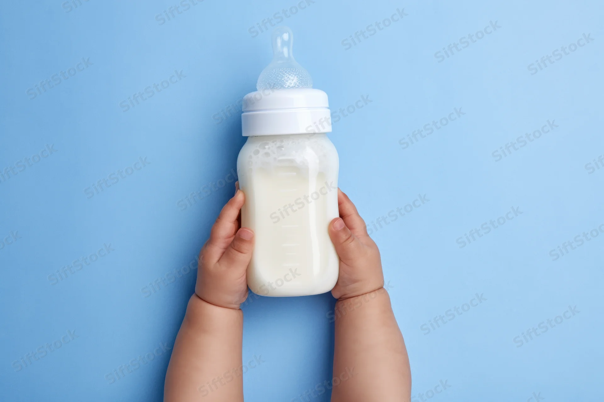 Baby hands holding a milk-filled bottle with measurement marks against a blue background