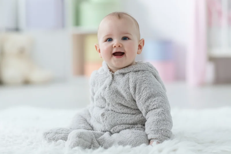 Baby in gray bunny onesie sitting on fluffy white rug smiling