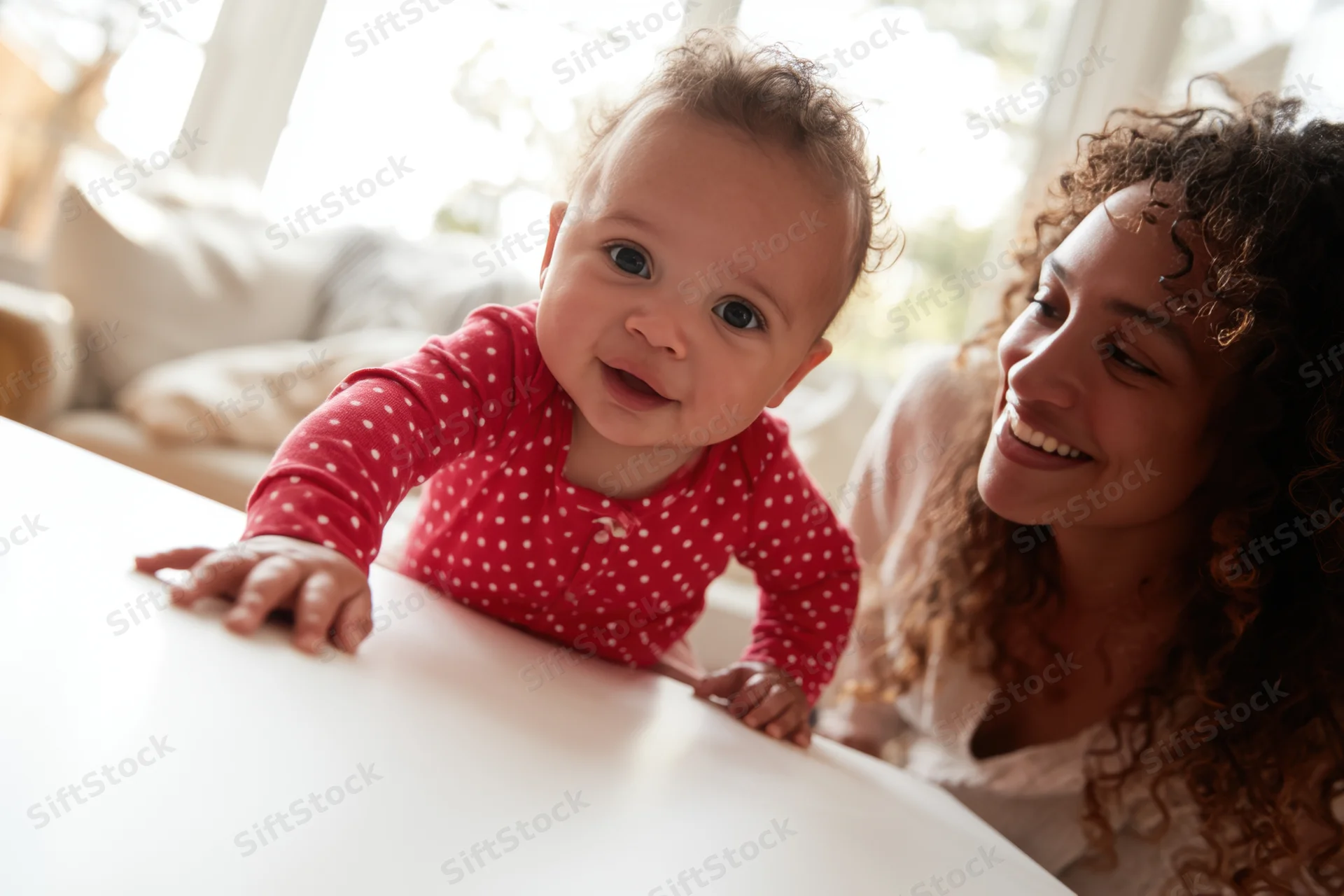 Baby in red polka-dot onesie reaching forward on white table with smiling mother beside, indoors near window