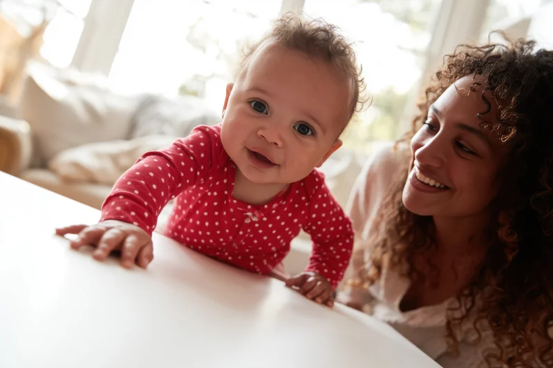 Baby in red polka-dot onesie reaching forward on white table with smiling mother beside, indoors near window