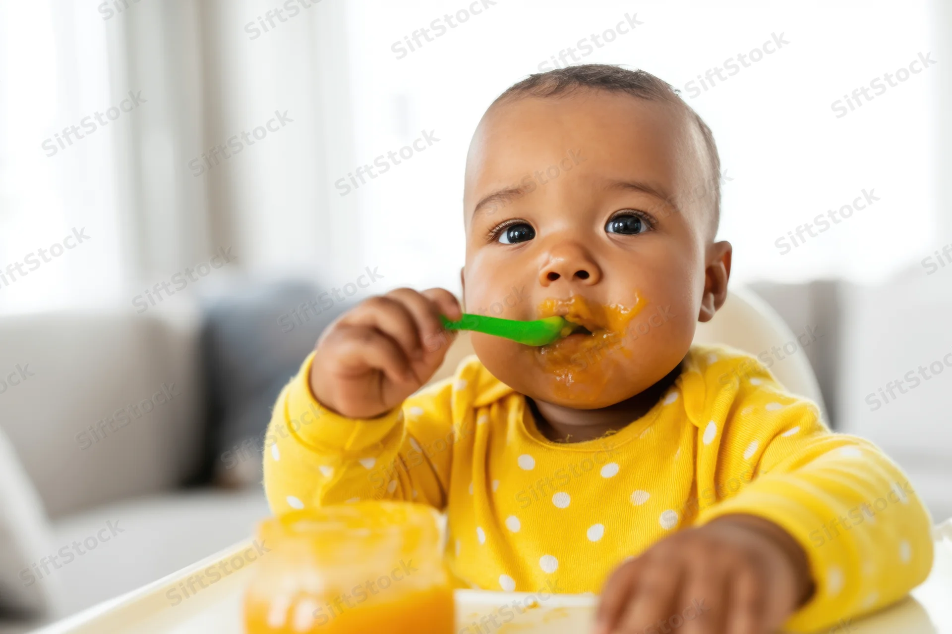 Baby in yellow polka-dot onesie eating puree with green spoon in high chair