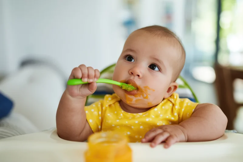 Baby in yellow polka-dot onesie eating puree with green spoon in high chair