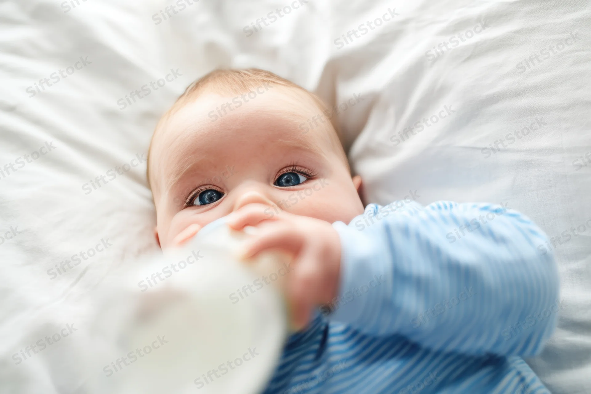 Baby lying on white pillow drinking from bottle in blue striped shirt