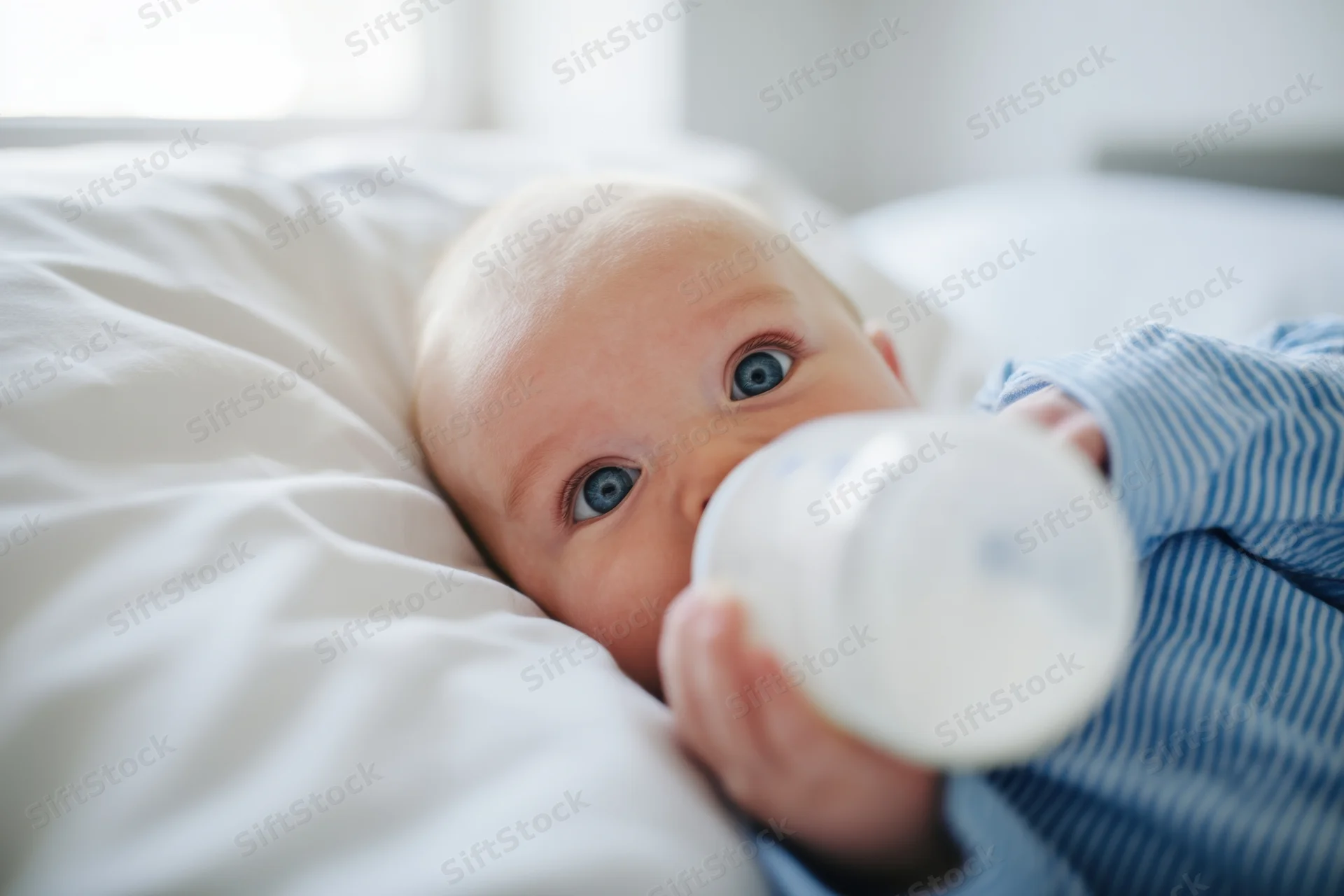Baby in blue striped shirt lying on white pillow drinking from bottle