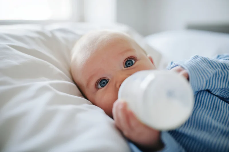 Baby in blue striped shirt lying on white pillow drinking from bottle