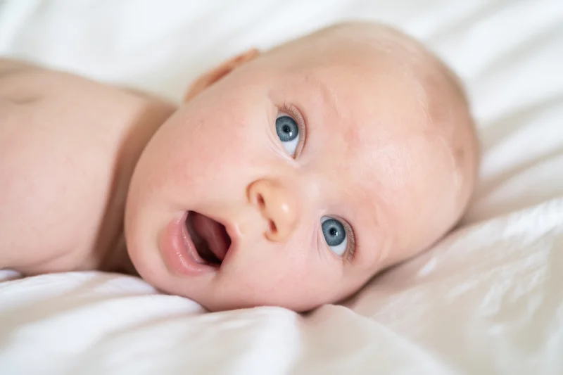 Baby lying on white bed with wide blue eyes and open mouth