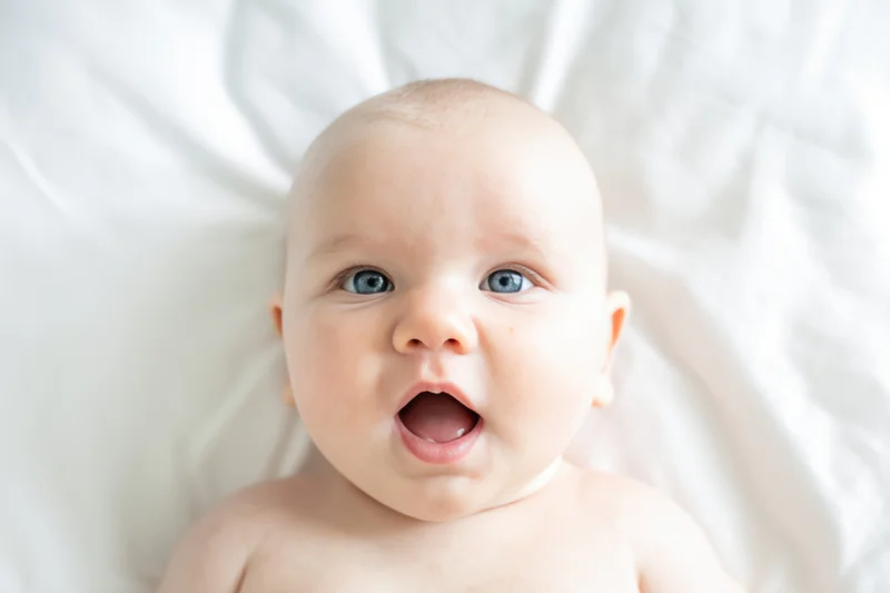 Baby lying on white bed with wide blue eyes and open mouth