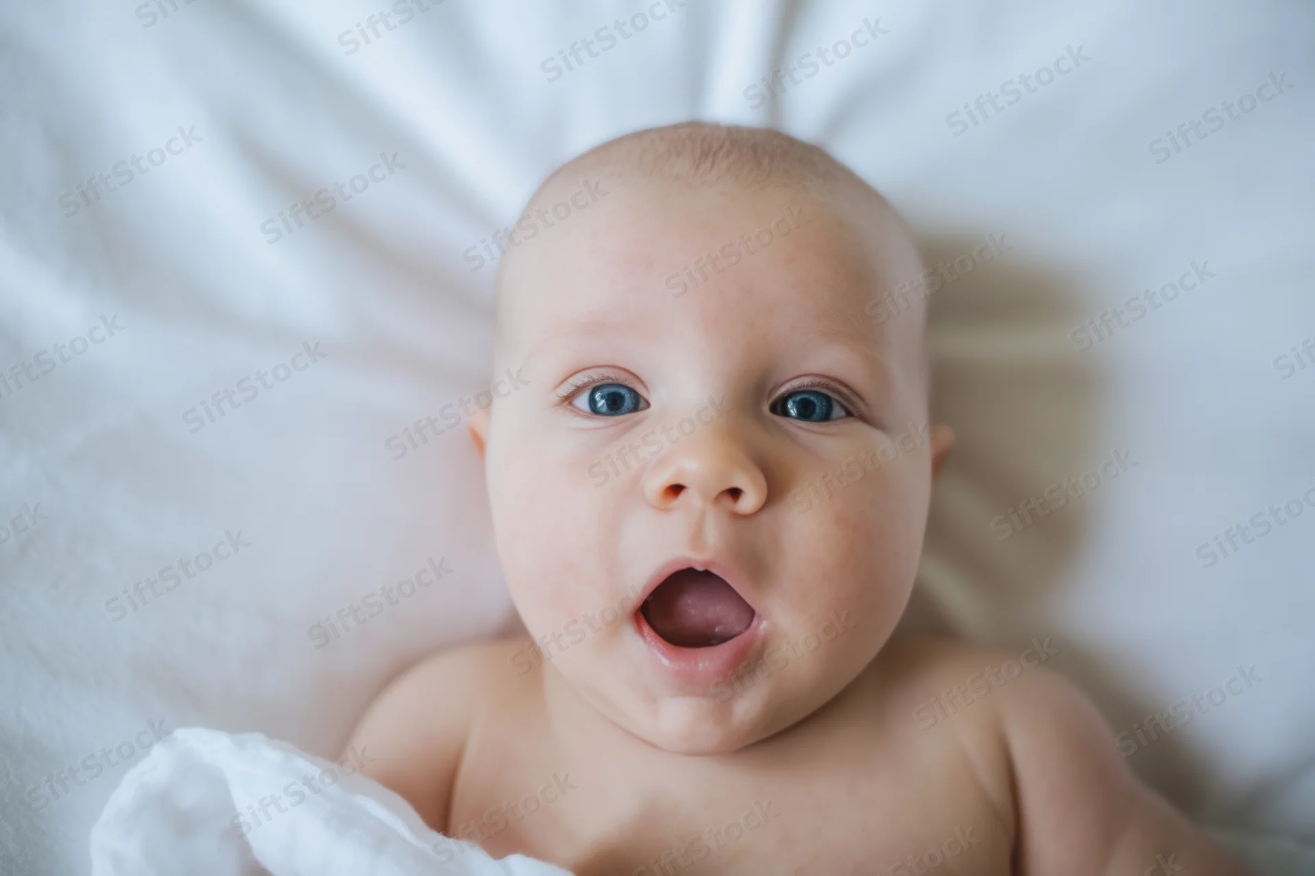 Baby lying on white bed with wide blue eyes and open mouth