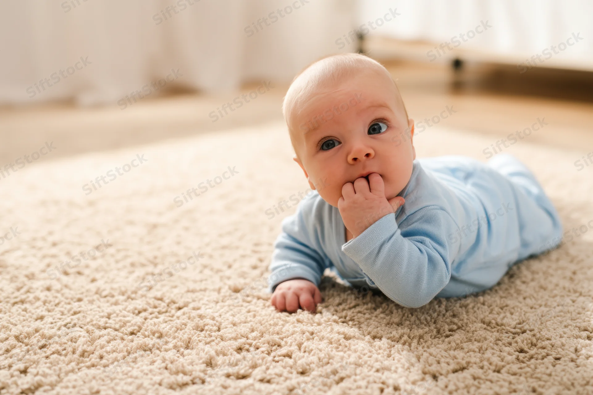 Baby lying on stomach on soft beige carpet in light blue onesie with hand in mouth