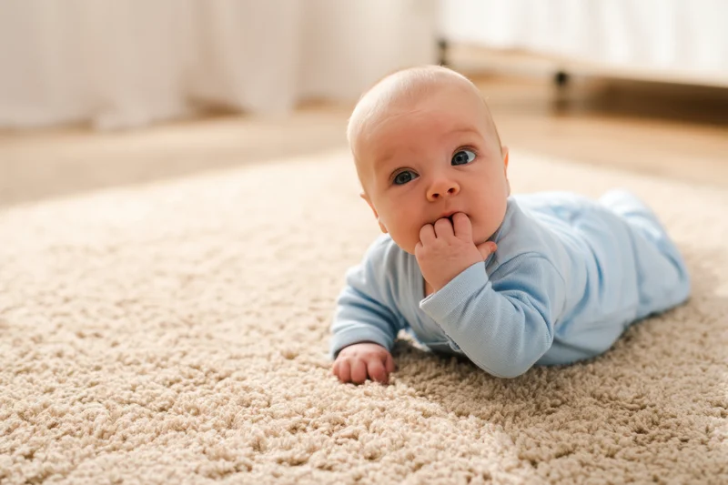 Baby lying on stomach on soft beige carpet in light blue onesie with hand in mouth