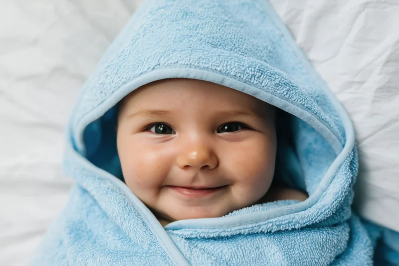 Baby wrapped in a light-blue hooded towel lying on a white bed, smiling at the camera