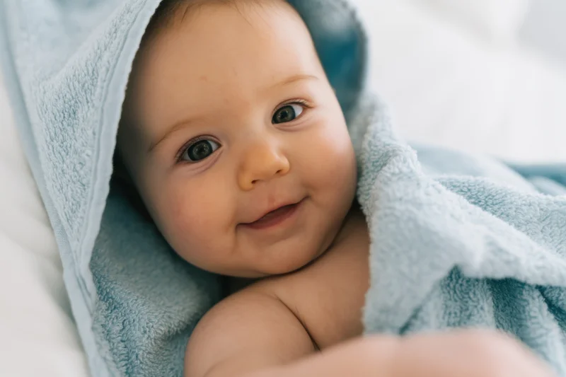 Baby wrapped in a soft blue hooded towel lying on a white bed smiling