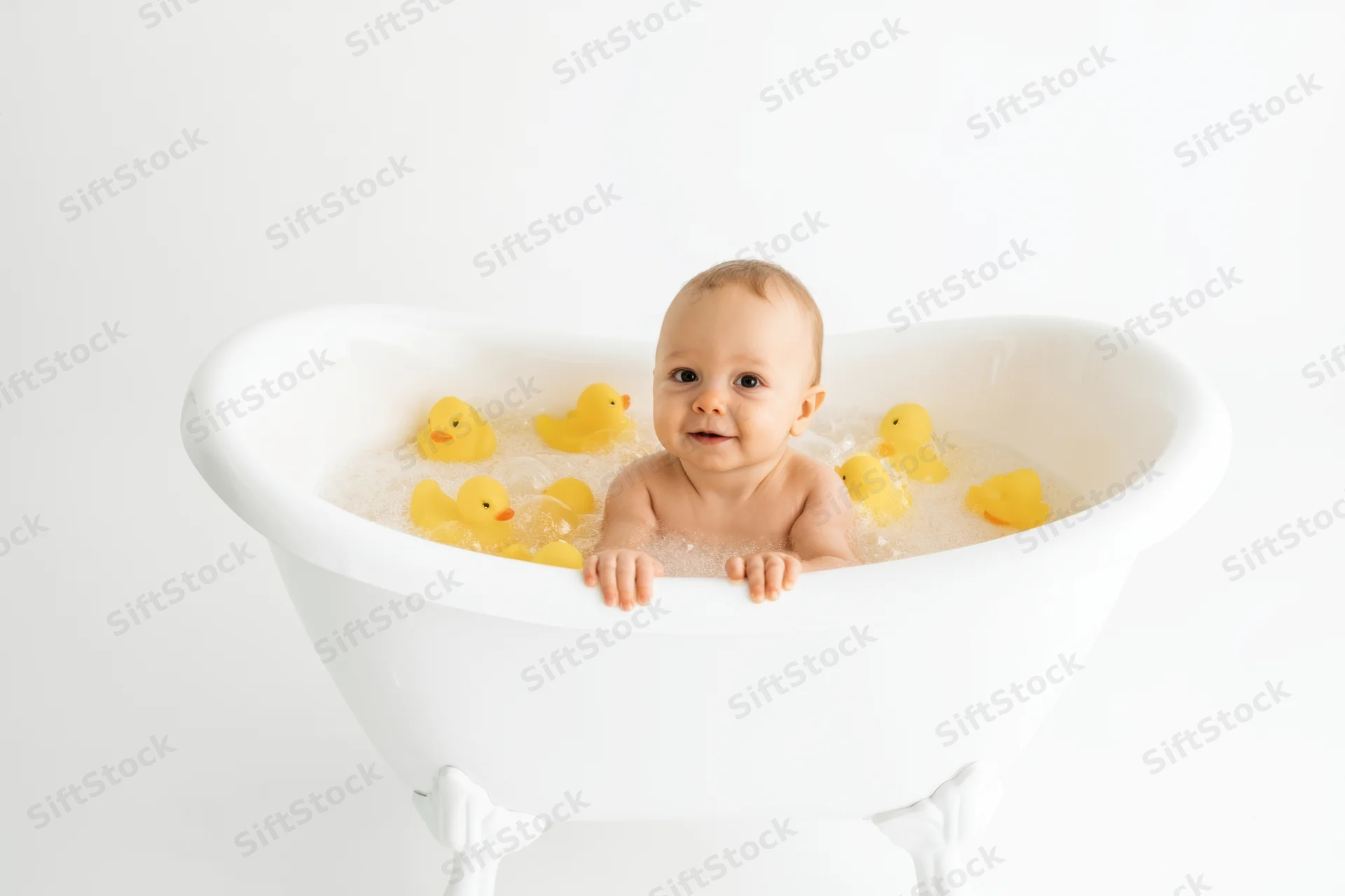 Baby sitting in a white clawfoot bathtub filled with water and yellow rubber ducks, smiling at the camera