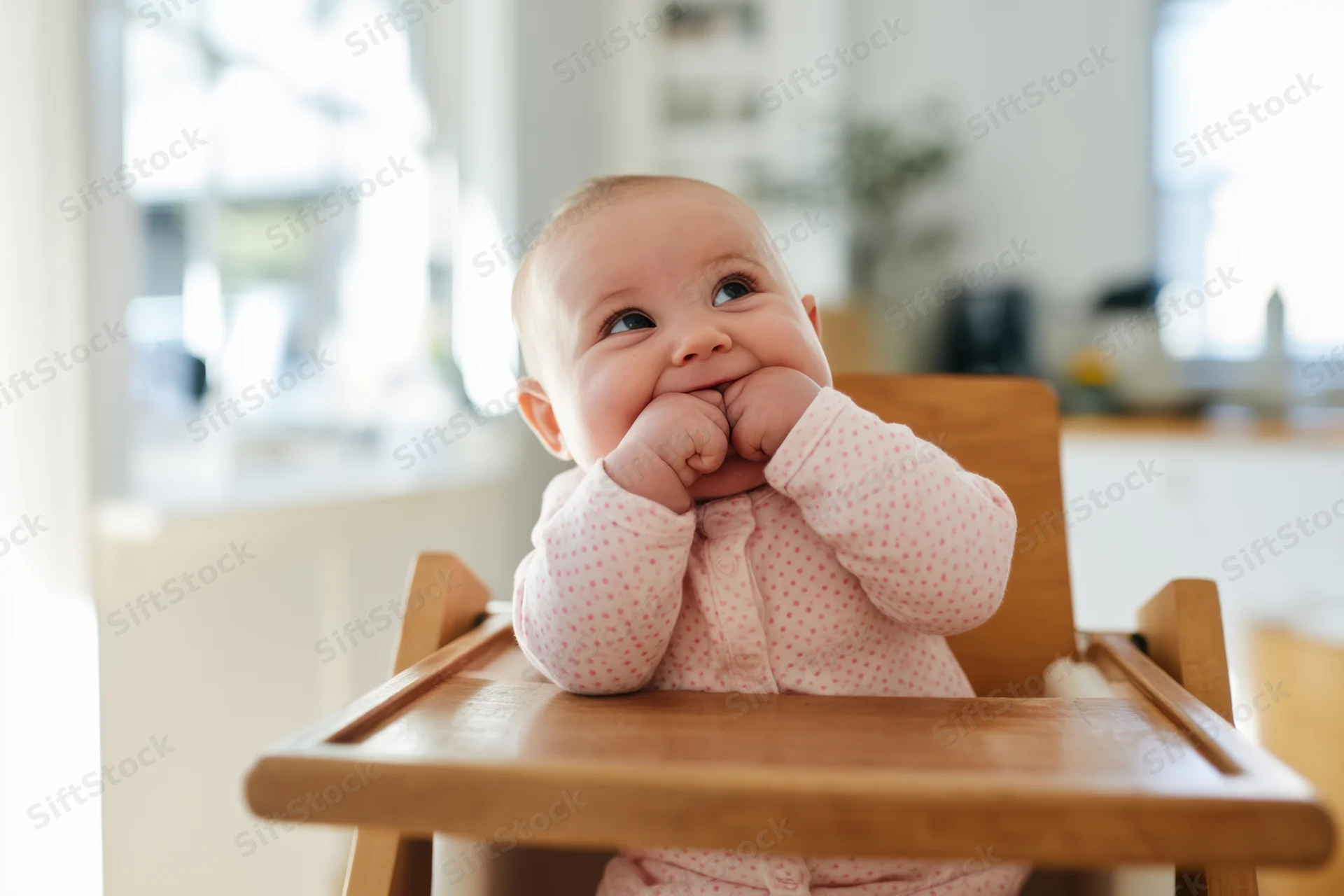 Baby sitting in wooden high chair chewing hands in bright kitchen